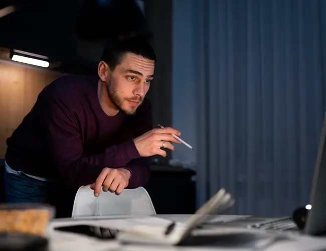 Man leaning over chair with pen in right hand while looking at laptop