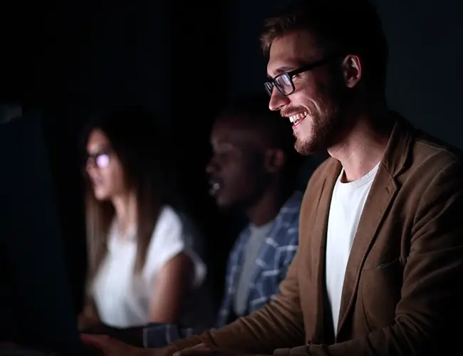 Man with glasses smiling while looking at laptop in dark room