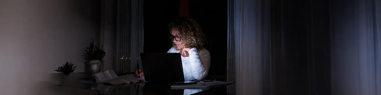 Woman with glasses sitting at desk working on laptop in dark room