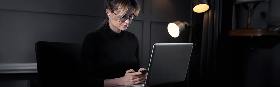 Woman with glasses on and earphones in typing on mobile phone with laptop in front