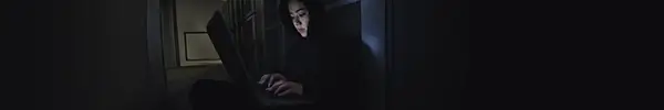 Woman with hood up typing on laptop in dark room