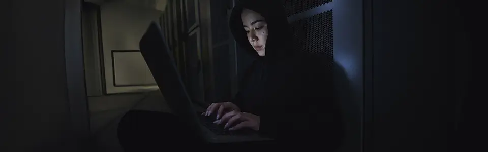 Woman with hood up typing on laptop in dark room