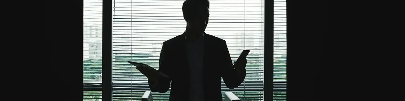 Silhouette of man standing in a dark room while holding a tablet and mobile phone