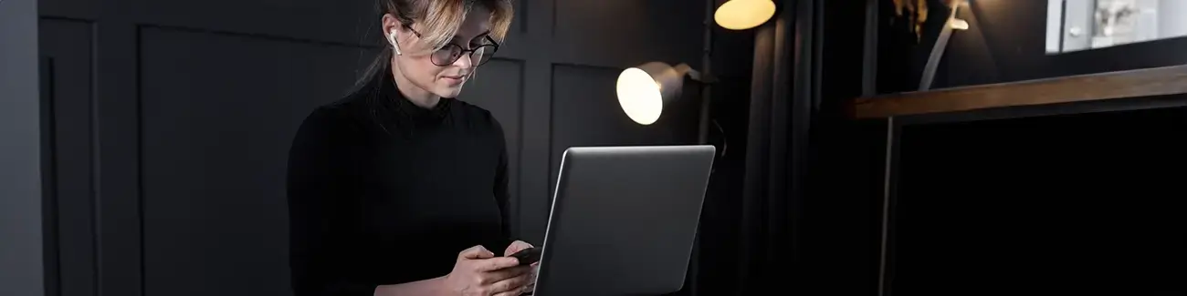 Woman with glasses on and earphones in typing on mobile phone with laptop in front