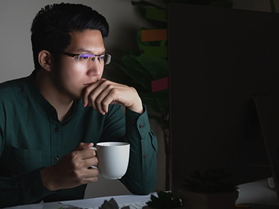 Man holding hand up to face with mug in right hand while looking at monitor in dark